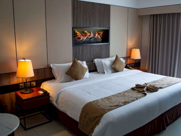 A hotel room features a large bed with bedside tables and lamps, wall art above the bed, a folded towel decoration, and a curtain-covered window.