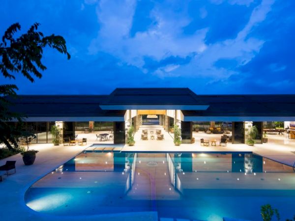 An illuminated outdoor pool area at dusk with a modern building in the background, featuring open seating spaces and lush surroundings.