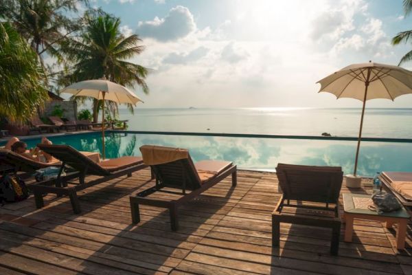 A serene poolside setting with wooden lounge chairs under umbrellas, overlooking a tranquil body of water and a scenic, sunny sky.