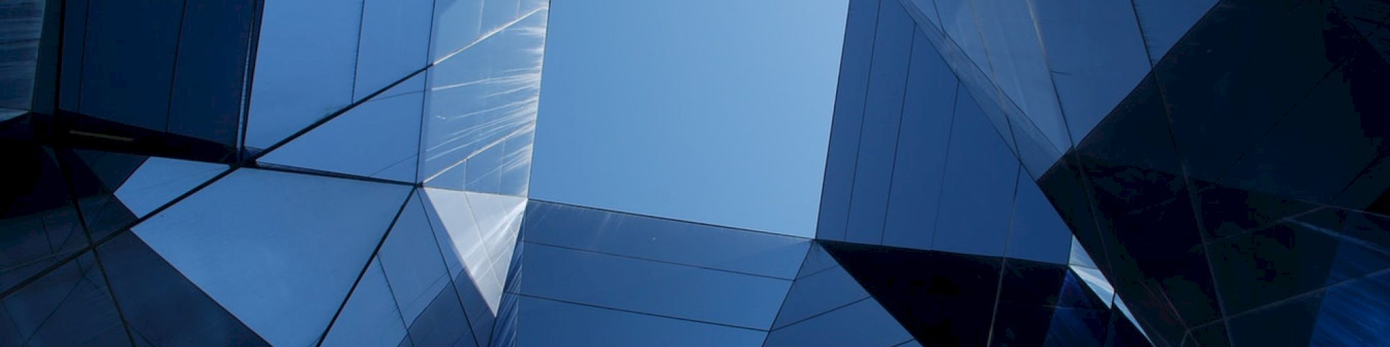 The image shows a geometric arrangement of reflective glass panels forming a tunnel-like structure with an opening at the top revealing the sky.