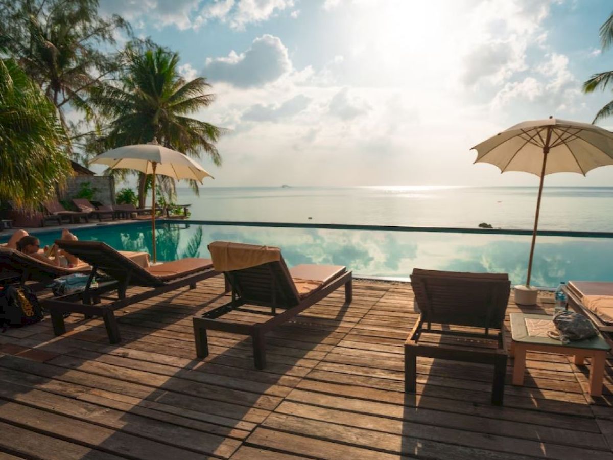 The image shows a serene poolside scene with lounge chairs, sun umbrellas, and a view of the ocean under a partly cloudy sky at sunrise or sunset.