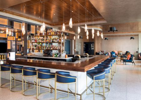 A modern bar with a wooden countertop, blue cushioned stools, pendant lights, and a well-stocked liquor shelf. There are seating areas in the background.