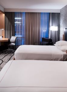 There is a modern hotel room with two twin beds, a wall-mounted TV, a work desk with a chair, and wall lamps on either side of the beds.