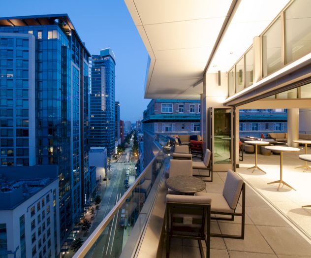 This image shows a modern cityscape at dusk with high-rise buildings and a rooftop terrace featuring tables, chairs, and a glass railing.