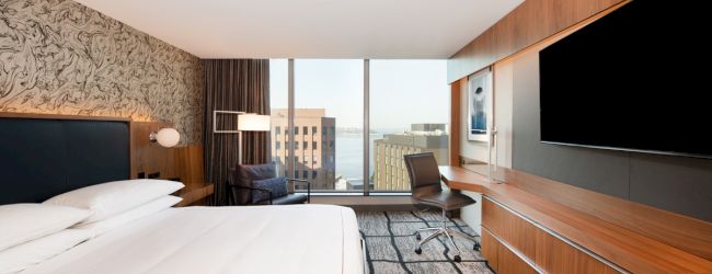 A modern hotel room featuring a king-sized bed, large TV, desk, chair, and window with a city view. The decor includes patterned walls and carpet.