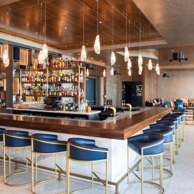 The image shows a modern bar with a wooden counter, blue barstools, hanging lights, and a variety of bottles. There is seating in the background.
