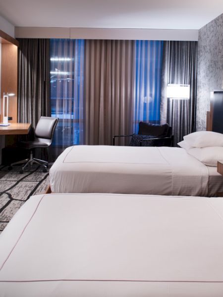 The image shows a modern hotel room with two double beds, a desk with a chair, bedside lamps, a flat-screen TV, and a large window with curtains.