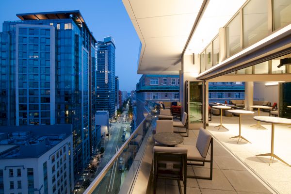 A modern urban balcony features outdoor seating and tables, with a view of high-rise buildings and a city street illuminated at dusk.