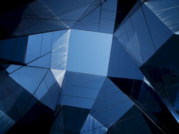 This is an abstract architectural image showing a view of the sky through mirrored, angular, glass-like structures, creating a geometric pattern.