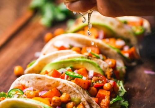 The image shows a close-up of five tacos with chickpeas, avocado, and vegetables, with a hand squeezing lime juice over them on a wooden surface.