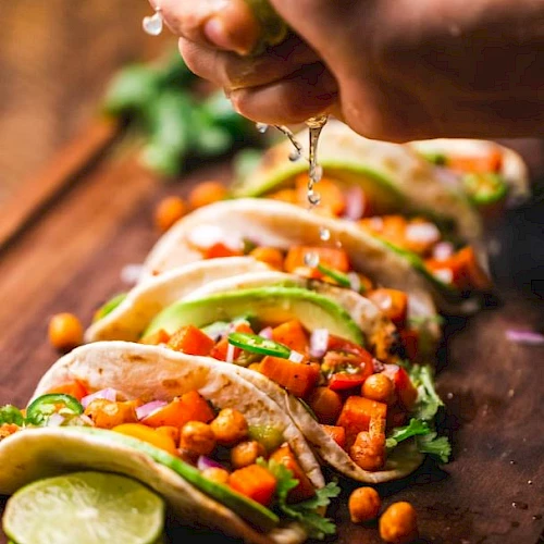 The image shows a close-up of five tacos with chickpeas, avocado, and vegetables, with a hand squeezing lime juice over them on a wooden surface.