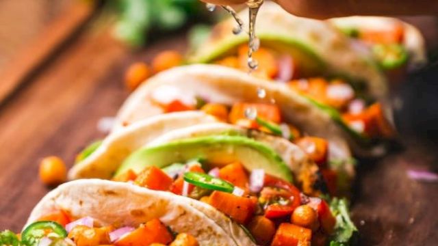 The image shows a close-up of five tacos with chickpeas, avocado, and vegetables, with a hand squeezing lime juice over them on a wooden surface.