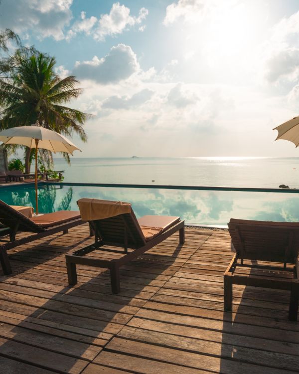 A serene tropical setting with lounge chairs, umbrellas, and an infinity pool overlooking a calm sea under a partly cloudy sky.