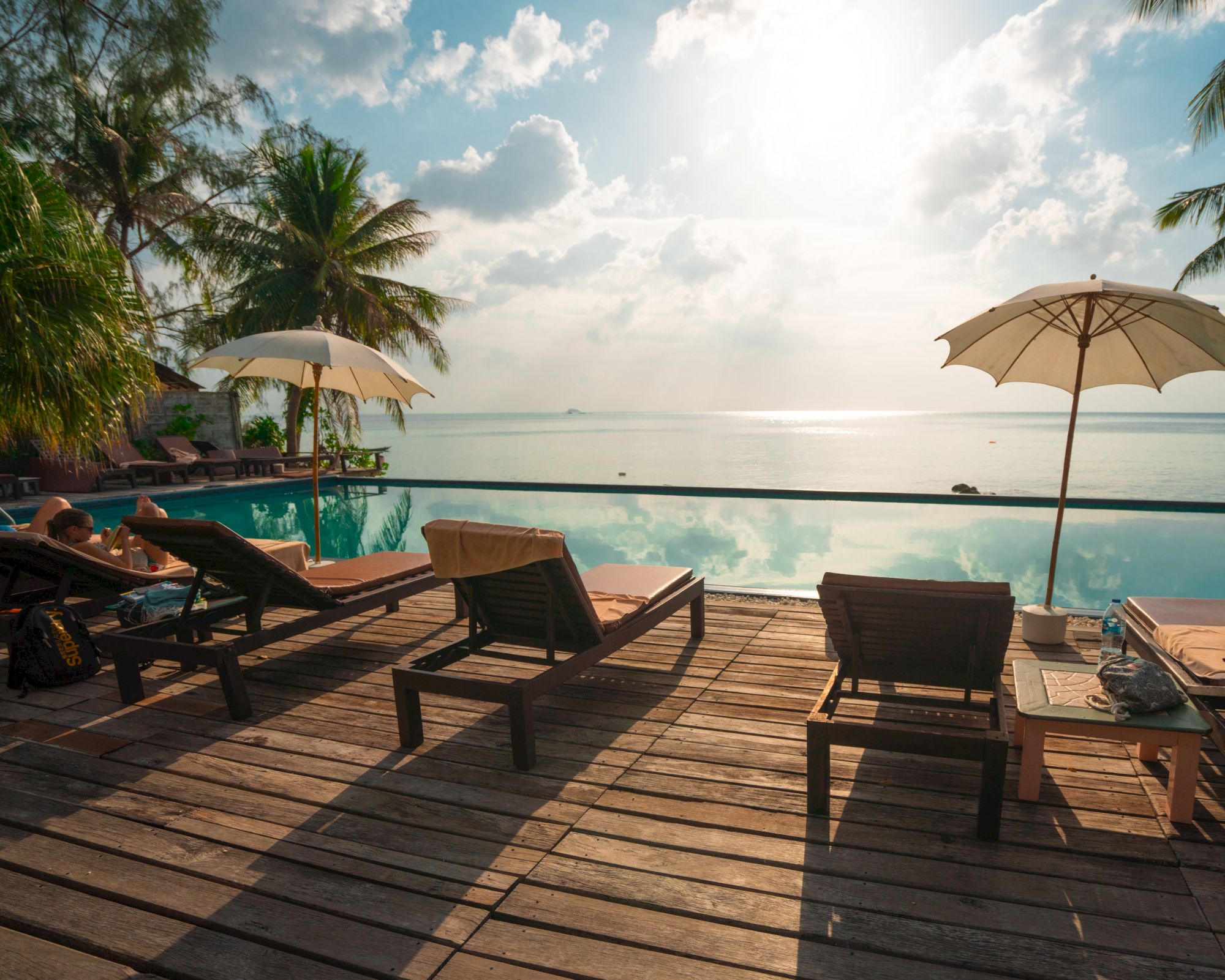 Sun loungers on a wooden deck by a pool, with umbrellas and a scenic view of the ocean under a partly cloudy sky, creating a relaxing atmosphere.