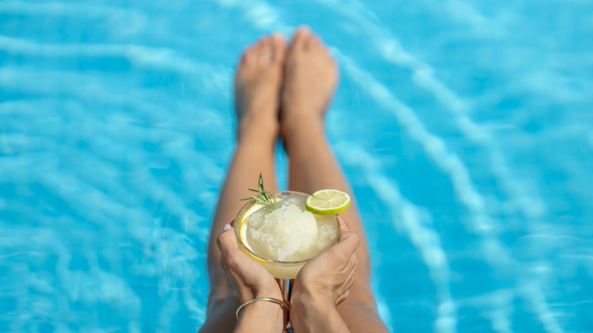 A person is relaxing by a pool with their legs in the water, holding a glass of a slushy drink topped with a lime slice and a sprig of rosemary.