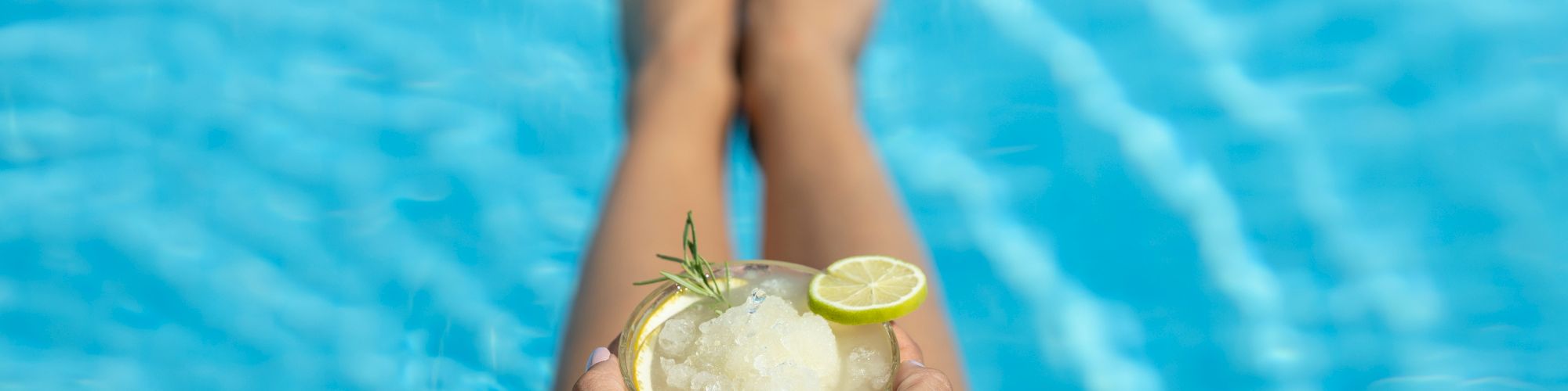 A person is relaxing by a pool with their legs in the water, holding a glass of a slushy drink topped with a lime slice and a sprig of rosemary.