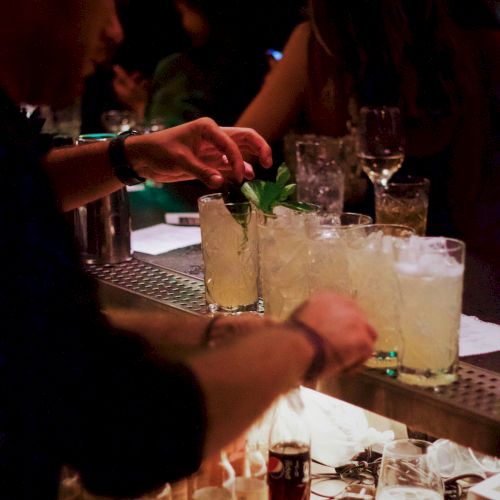 A bartender is preparing cocktails at a bar, topping them with mint garnish. Various bottles and glasses are visible on the counter in a dimly lit setting.