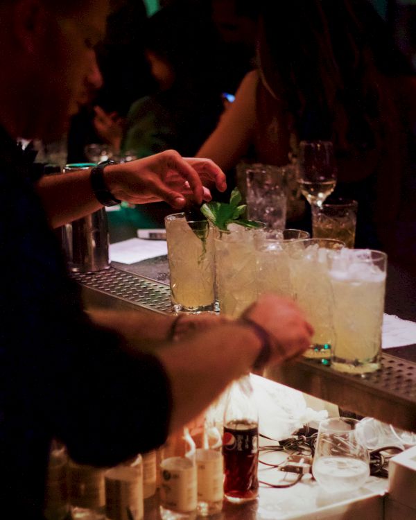 A bartender is preparing cocktails at a bar, topping them with mint garnish. Various bottles and glasses are visible on the counter in a dimly lit setting.