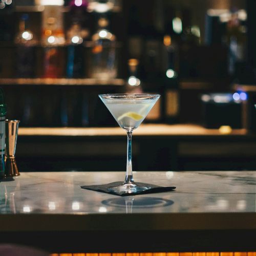 A martini glass with a lemon twist sits on a marble bar counter in a dimly lit bar with bottles and soft lighting in the background.