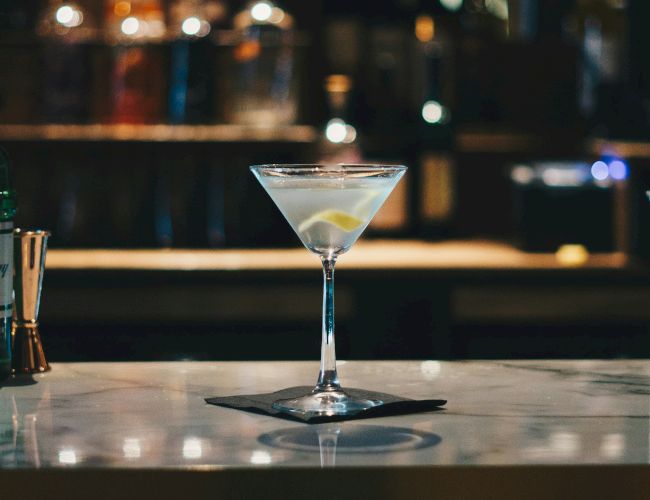 A martini glass with a lemon twist sits on a marble bar counter in a dimly lit bar with bottles and soft lighting in the background.