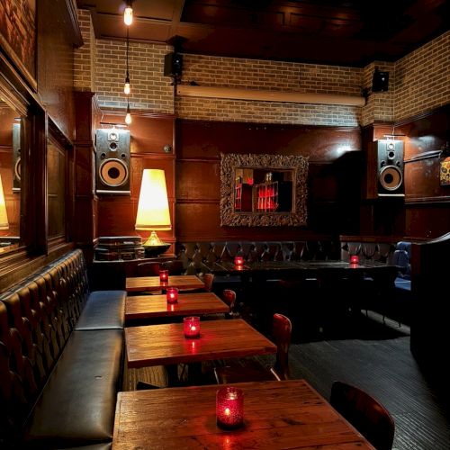 A dimly lit bar interior with wooden tables, red candles, leather seating, and a warm ambiance.