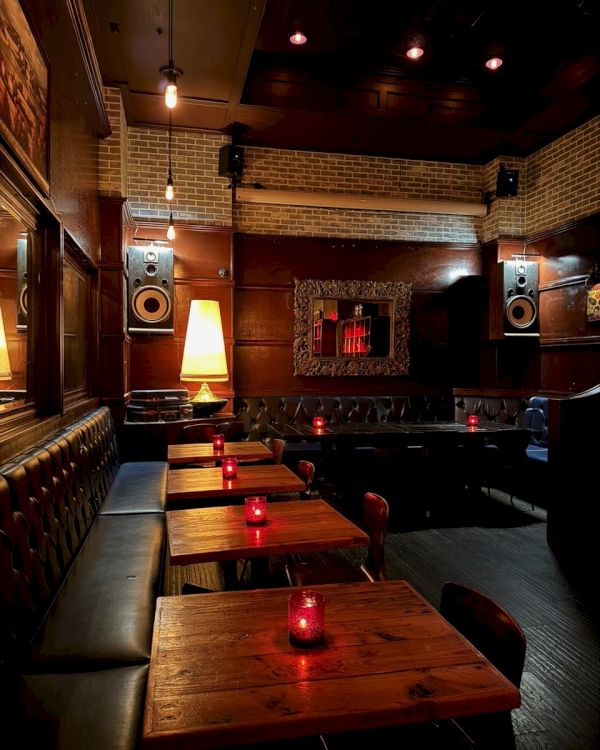 A dimly lit bar interior with wooden tables, red candles, leather seating, and a warm ambiance.