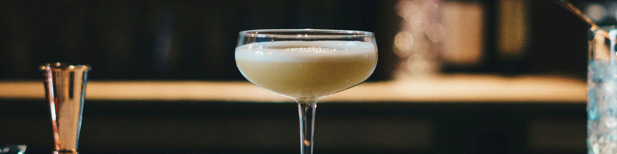 A cocktail glass filled with a creamy white drink is placed on a dimly lit bar counter, with various bottles and bar tools in the background.