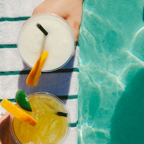 Two hands are holding drinks with garnishes over a striped towel by a pool. The drinks appear to be a yellow cocktail and a white smoothie.