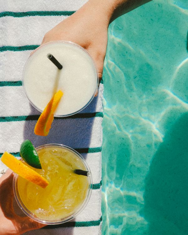 Two hands are holding drinks with garnishes over a striped towel by a pool. The drinks appear to be a yellow cocktail and a white smoothie.