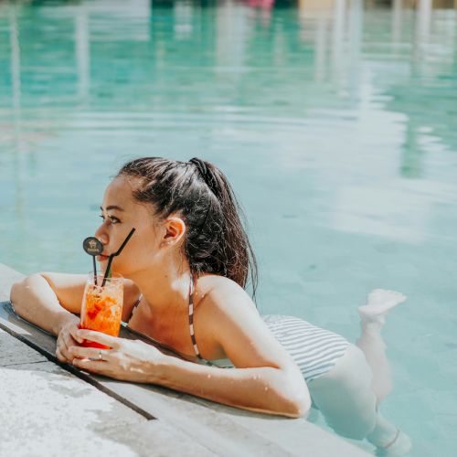 A woman is relaxing in a swimming pool, holding a drink with a straw and a garnish, while leaning on the pool edge, gazing into the distance.