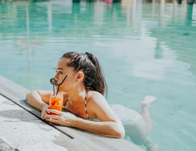 A woman is relaxing in a swimming pool, holding a drink with a straw and a garnish, while leaning on the pool edge, gazing into the distance.