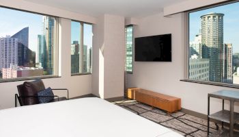 This image shows a modern, hotel room with large windows offering a cityscape view, a wall-mounted TV, an armchair, and a bed in the foreground.