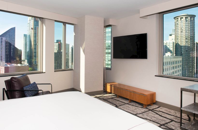 This image shows a modern, hotel room with large windows offering a cityscape view, a wall-mounted TV, an armchair, and a bed in the foreground.