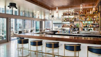 This image shows a modern bar with a wooden counter, blue and gold barstools, and a wide selection of liquor bottles on shelves.