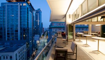 A modern outdoor terrace overlooks a city street with tall buildings at dusk, featuring tables, chairs, and a bar area under warm lighting.