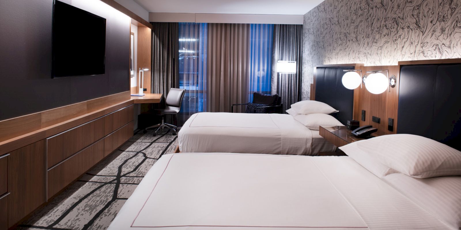 A modern hotel room with two double beds, a wall-mounted TV, a desk with a chair, and contemporary decor including curtains and lamps.
