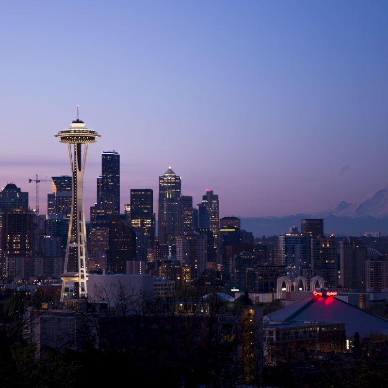 This image depicts the Seattle skyline at dusk, with the iconic Space Needle prominently visible and Mount Rainier in the background.