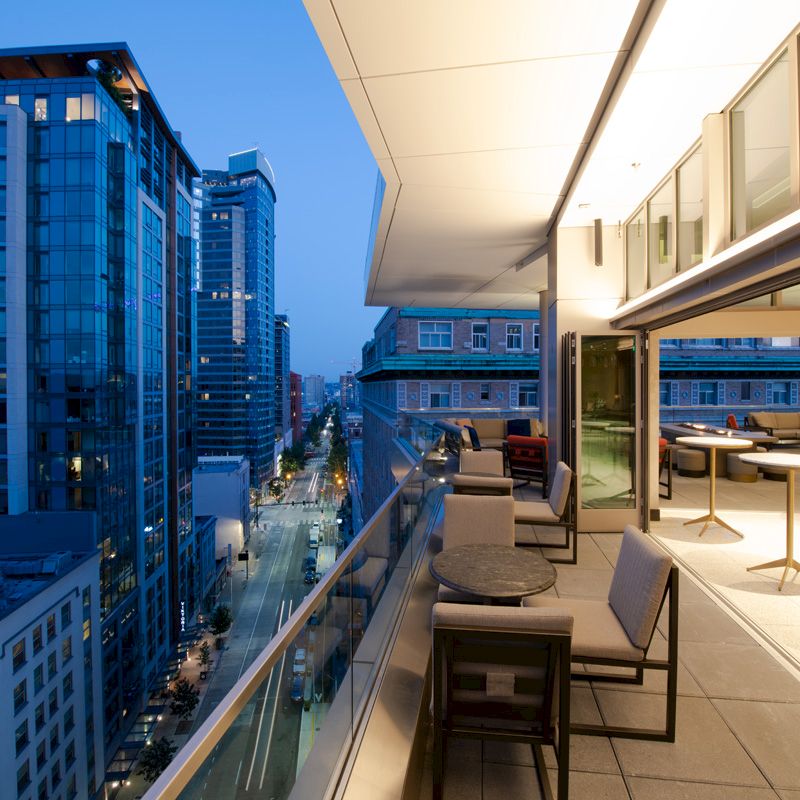 A modern cityscape at dusk with high-rise buildings and a stylish balcony featuring outdoor seating and tables, overlooking the urban scene.