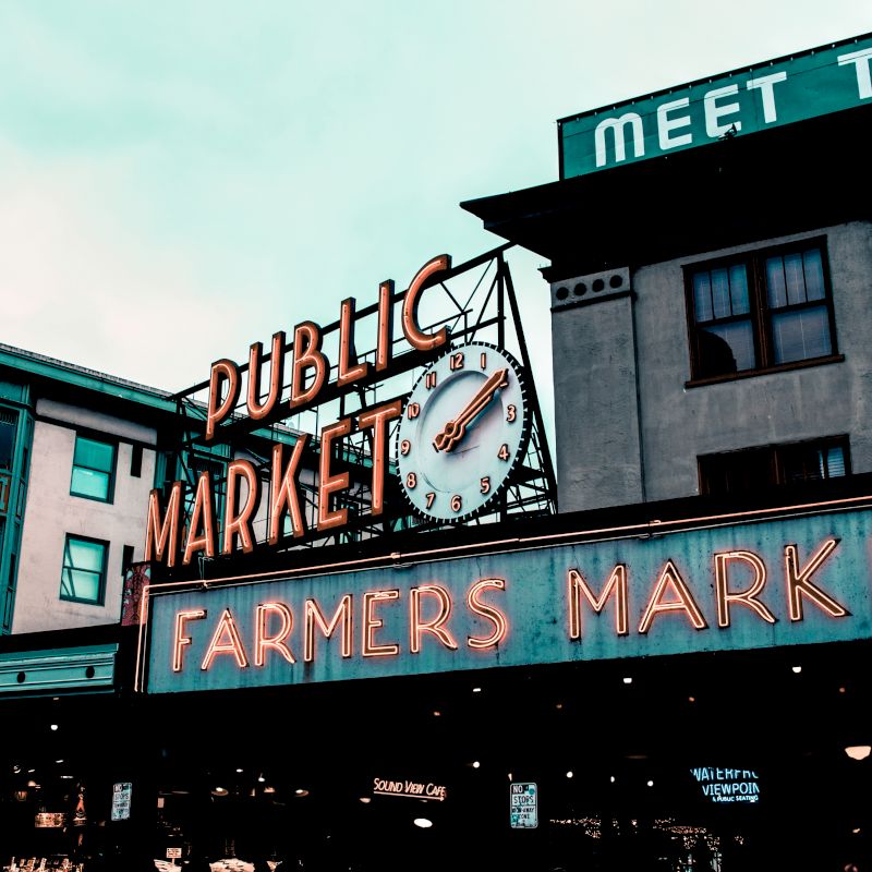Image depicts the vibrant entrance of a farmers market with neon signs, a clock, and adjacent buildings, creating a bustling urban atmosphere.