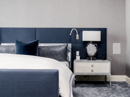 A modern bedroom featuring a bed with blue upholstery, a white nightstand, a decorative lamp, and a gray carpet.