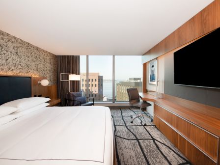 A modern hotel room with a large bed, wall-mounted TV, desk, chair, and a window view of buildings and water is visible.