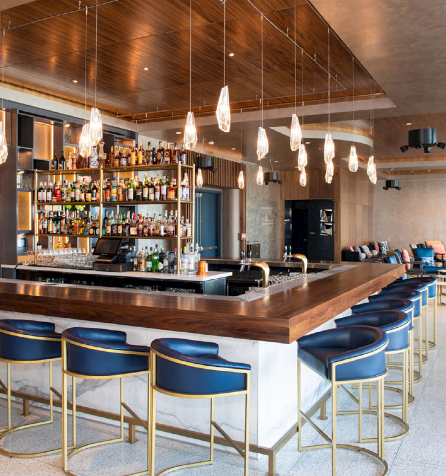 The image shows a modern bar with a wooden counter, blue bar stools, and stylish lighting fixtures. Shelves behind hold various bottles of liquor.