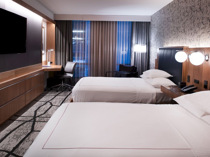 A modern hotel room with two neatly made double beds, a flat-screen TV, a work desk, a chair, and a window with curtains and a view.