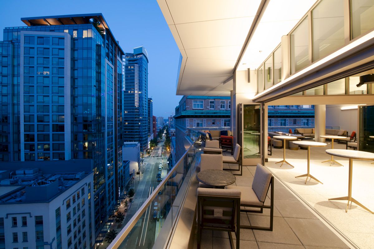 A rooftop terrace with tables and chairs overlooks a cityscape with tall buildings and a street lit up in the evening.