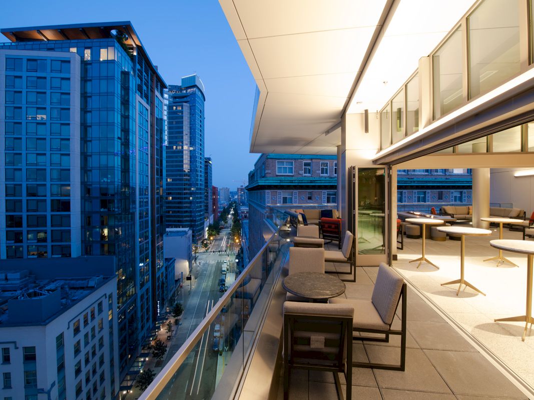 A rooftop terrace with tables and chairs overlooks a cityscape with tall buildings and a street lit up in the evening.