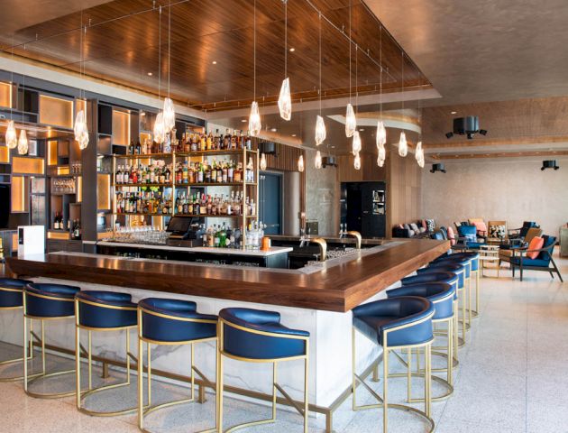 A modern bar with a central counter, rows of blue and gold bar stools, shelves stocked with bottles, pendant lights, and a seating area in the background.