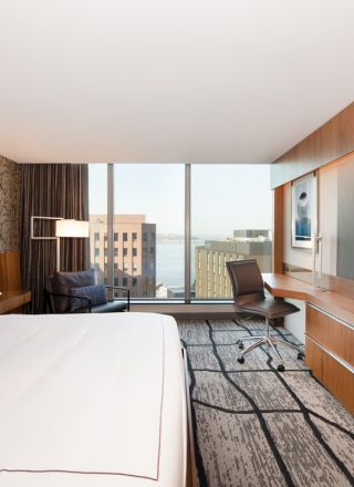 The image shows a modern hotel room with a large bed, a desk, a chair, a flat-screen TV, and a window with a city view and buildings in the distance.