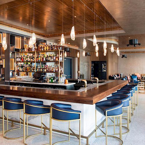 A modern bar with a wooden counter, blue barstools, and hanging lights. Shelves behind the bar are stocked with various bottles.