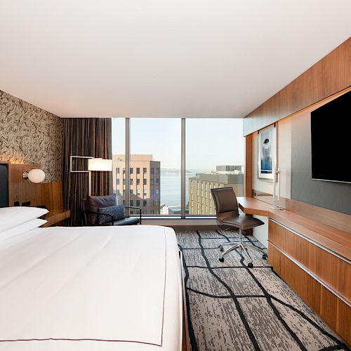 A modern hotel room with a large bed, wall-mounted TV, desk, chair, and floor-to-ceiling window overlooking a city and water view, is shown.