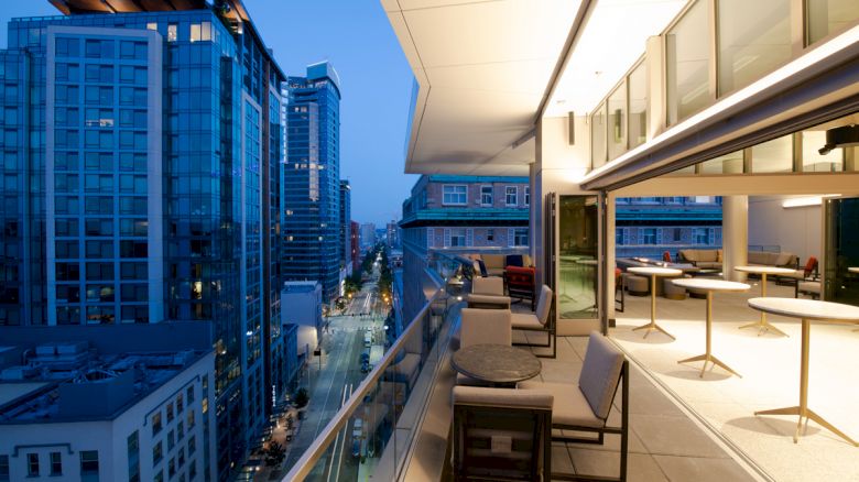 A modern cityscape with tall buildings at dusk, featuring an outdoor terrace and seating areas with a view of the street below.